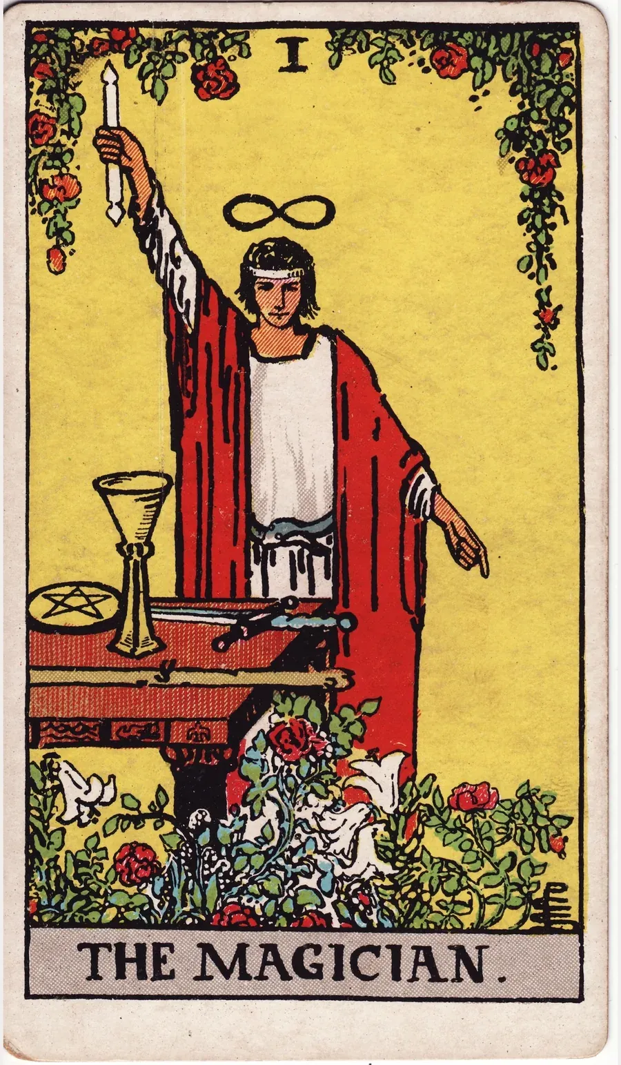 A young man in a red robe over white raises a wand toward the sky and points his other hand to the ground, with a cup, sword, pentacle and wand on the table before him